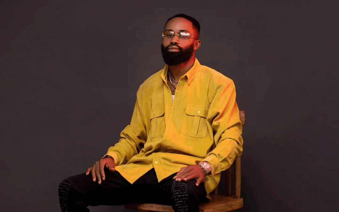 A man with a full beard and glasses sits confidently on a wooden chair against a dark background. He wears a loose-fitting yellow button-up shirt and black pants, accessorized with a wristwatch, rings, and necklaces. Warm light highlights his left side.