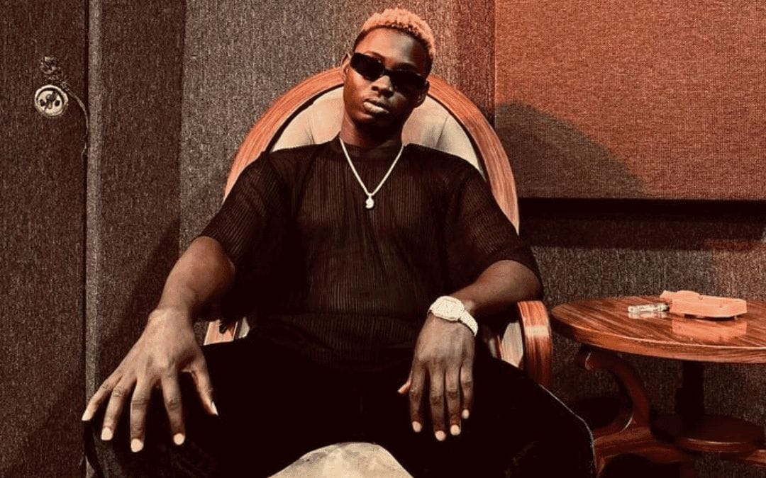 A man with short blond hair sits confidently in a wooden armchair, wearing dark sunglasses, a sheer black shirt, black pants, a watch, and a chain necklace. He poses with arms resting on the chair, in a room with brown soundproof walls.