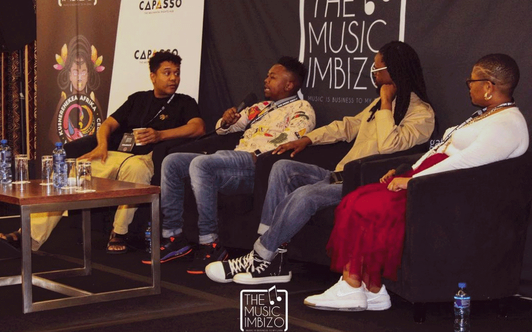 Four people sit on stage in armchairs, engaged in a panel discussion at The Music Imbizo event. They hold microphones and water bottles rest on a nearby table. Event banners and logos are visible in the background, creating a professional conference setting.