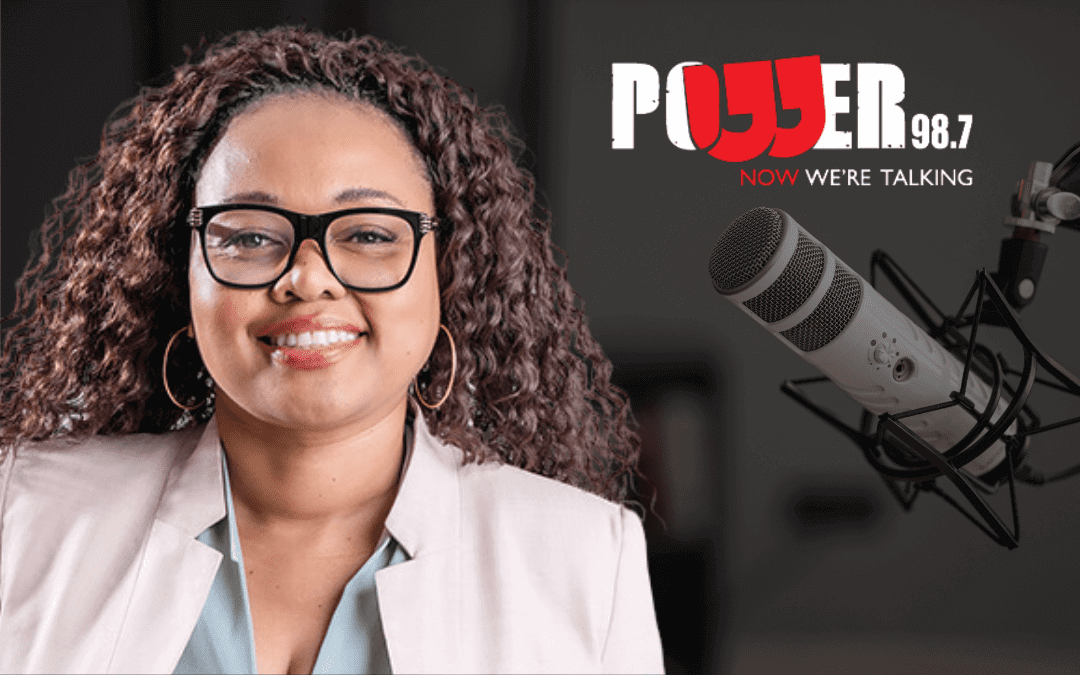 A smiling woman with curly hair and glasses wears a light-colored blazer. Beside her is a large microphone. In the background, text reads “POWER 98.7 NOW WE'RE TALKING” with “POWER” in bold, white and red letters. The setting appears to be a radio studio.