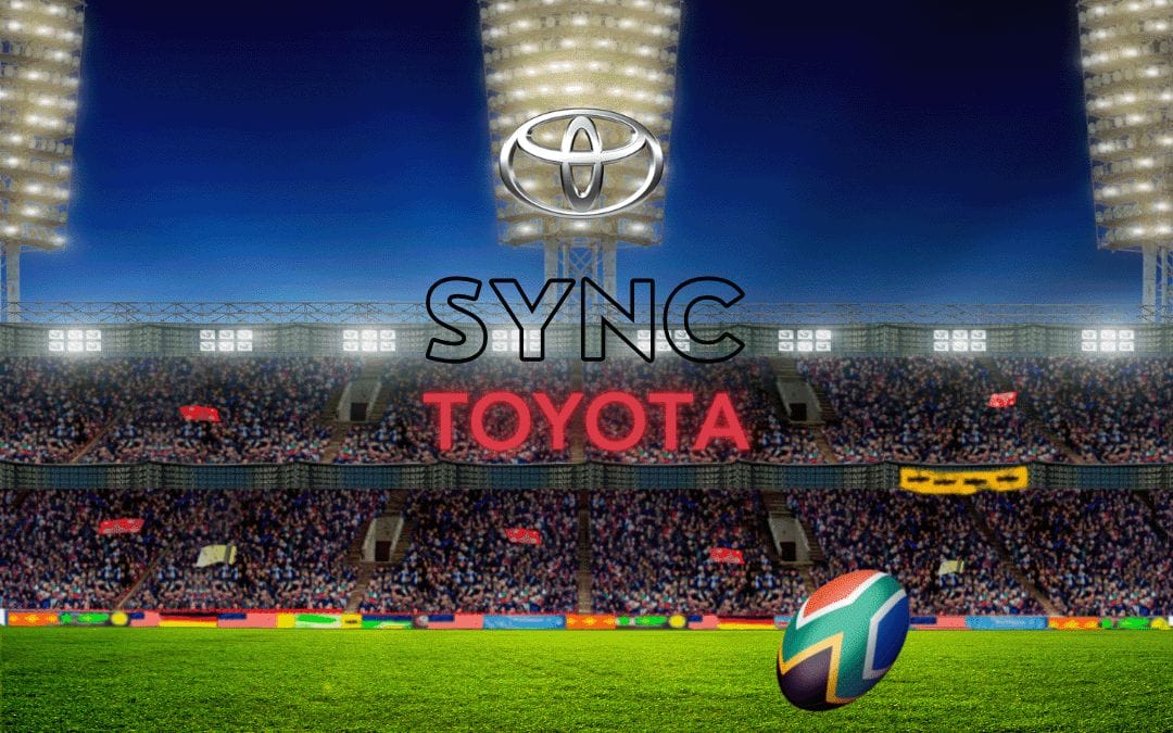 A rugby stadium under bright lights is packed with fans. Centered, a Toyota logo hovers above the words “SYNC” and “TOYOTA.” A rugby ball, marked with South African flag colors, rests on the green field in the foreground.