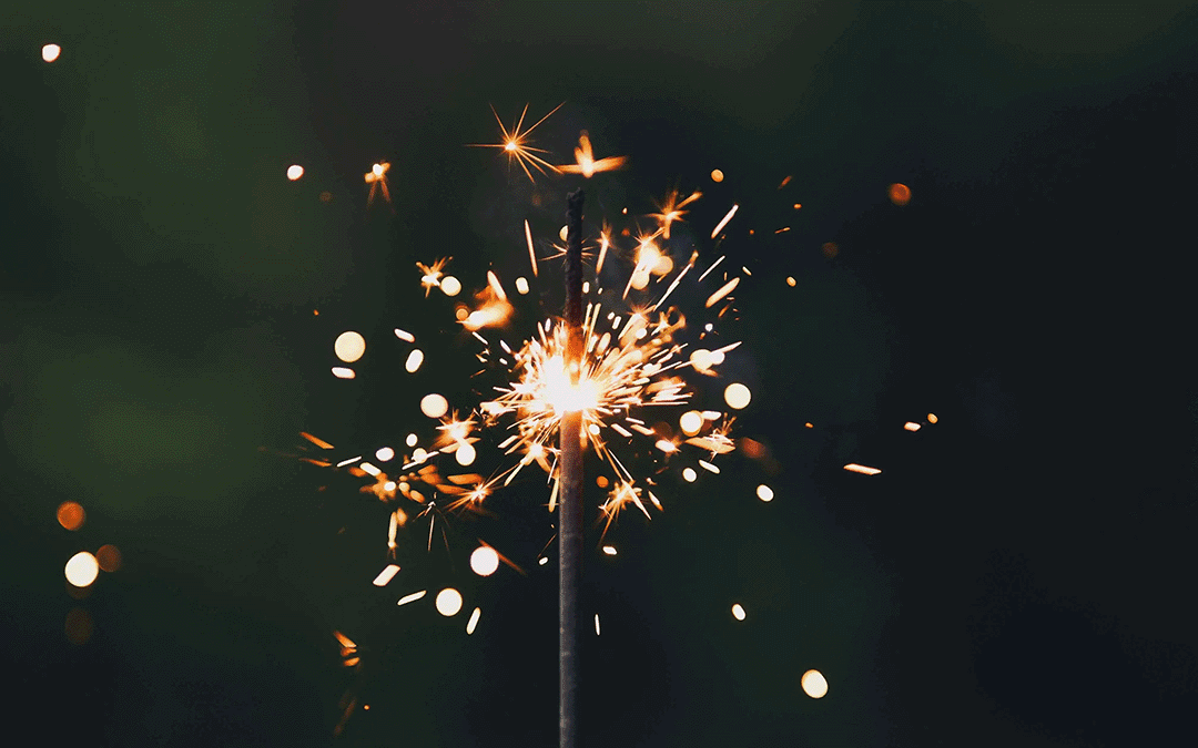 A close-up of a lit sparkler against a dark, blurred background. Bright orange and white sparks radiate outward from the tip, creating dynamic bursts of light, with glowing embers and fine trails trailing away from the centre.