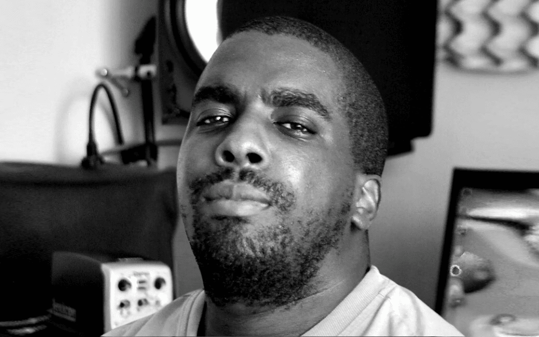 A man with a short beard and moustache looks confidently at the camera. He is in a room with audio equipment and a patterned object visible in the background. The image is in black and white.