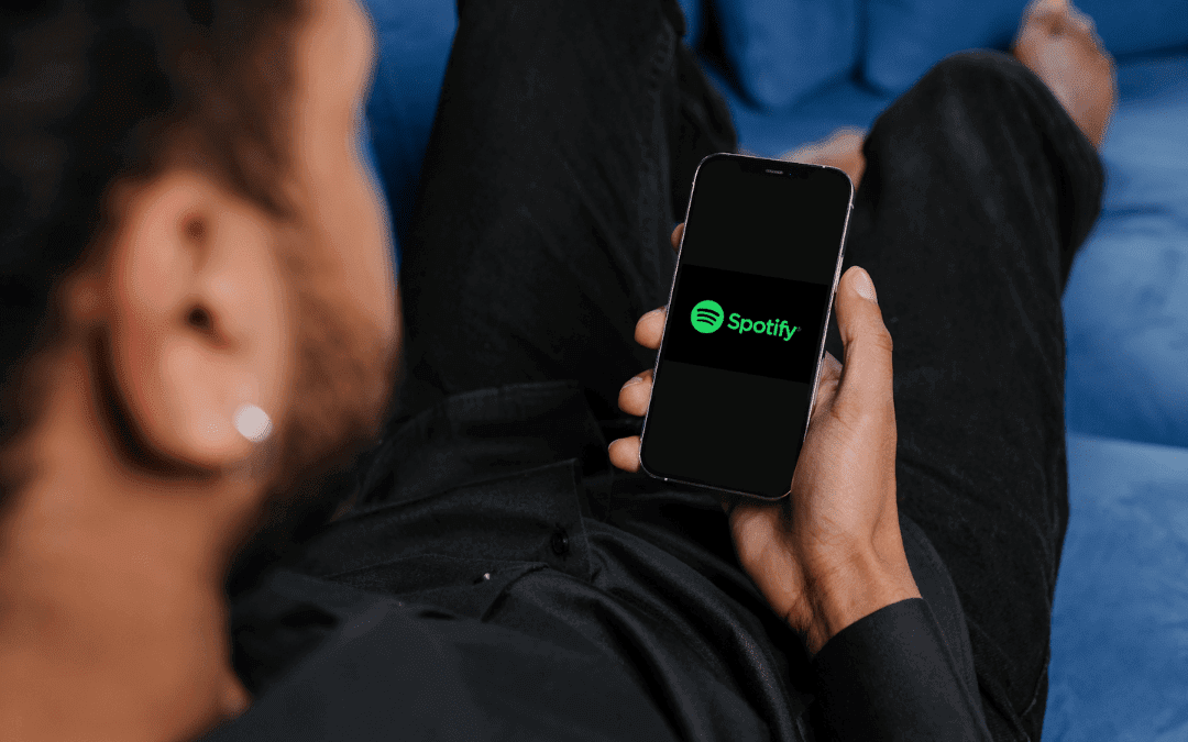 A person with short, dark hair and an earring relaxes on a blue sofa, holding a mobile phone in one hand. The phone screen displays the Spotify app logo on a black background. The individual wears a black shirt and is viewed from behind and slightly to the side.