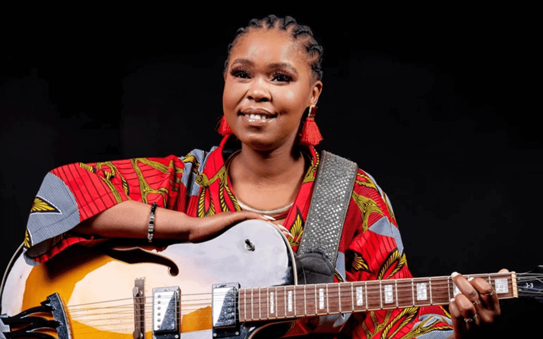 A smiling woman with plaited hair and red earrings wears a colourful, patterned shirt. She is holding an acoustic guitar, with her left hand on the fretboard and her right arm resting on the body. The background is dark, making her and the guitar stand out.