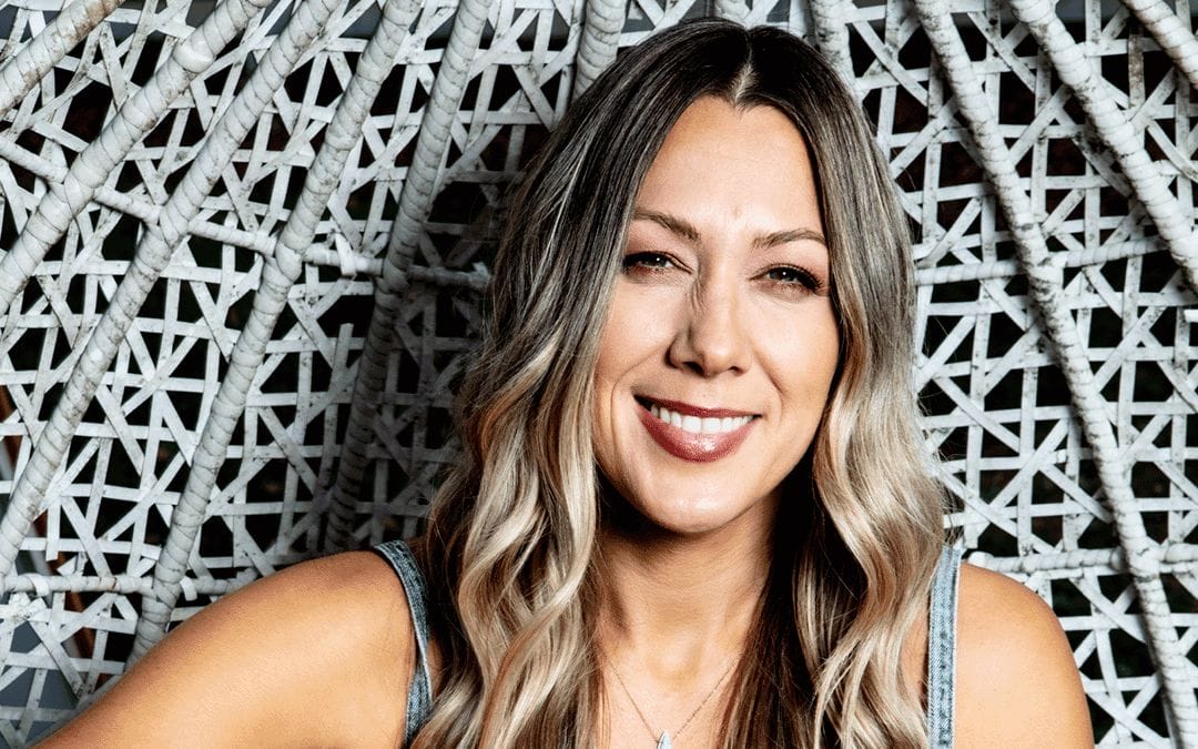 A woman with long, wavy, blonde and brown hair smiles at the camera. She is sitting in front of a white, woven, lattice-style chair. She wears a denim-style sleeveless top and a necklace. The background is slightly blurred.