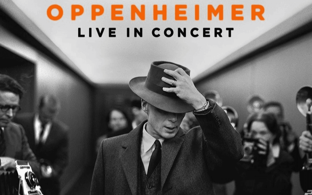 Black-and-white image of a man in a suit and hat, tilting his hat downward whilst walking through a corridor lined with photographers and reporters. Above, orange and black text reads: “OPPENHEIMER LIVE IN CONCERT.”.