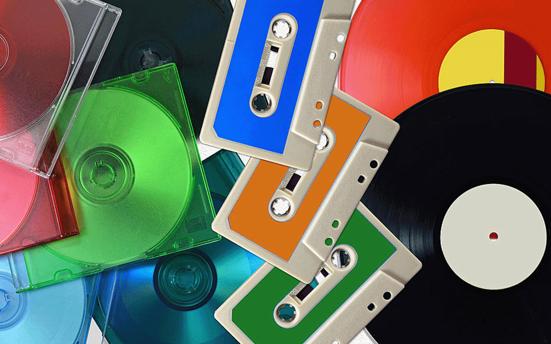 A colourful assortment of music formats: several translucent CDs in green, blue, and red cases, three audio cassettes in blue, orange, and green, and parts of two vinyl records, one black and one red, overlapping each other.