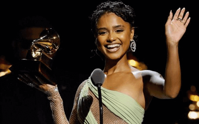 A woman in a green, one-shoulder gown smiles and waves whilst holding a Grammy award. She stands in front of a microphone on stage, with dramatic lighting highlighting her and the golden trophy.