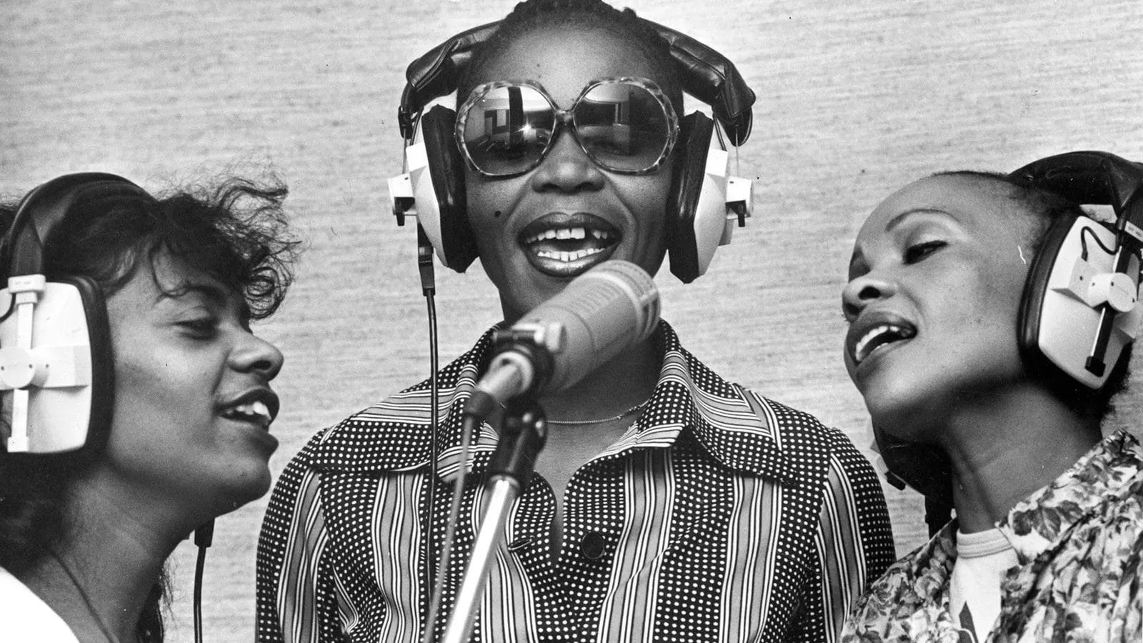 Three women wearing large headphones sing closely together into a studio microphone. The woman in the centre wears sunglasses and a striped shirt. All three appear joyful and focused, captured in black and white against a plain background.