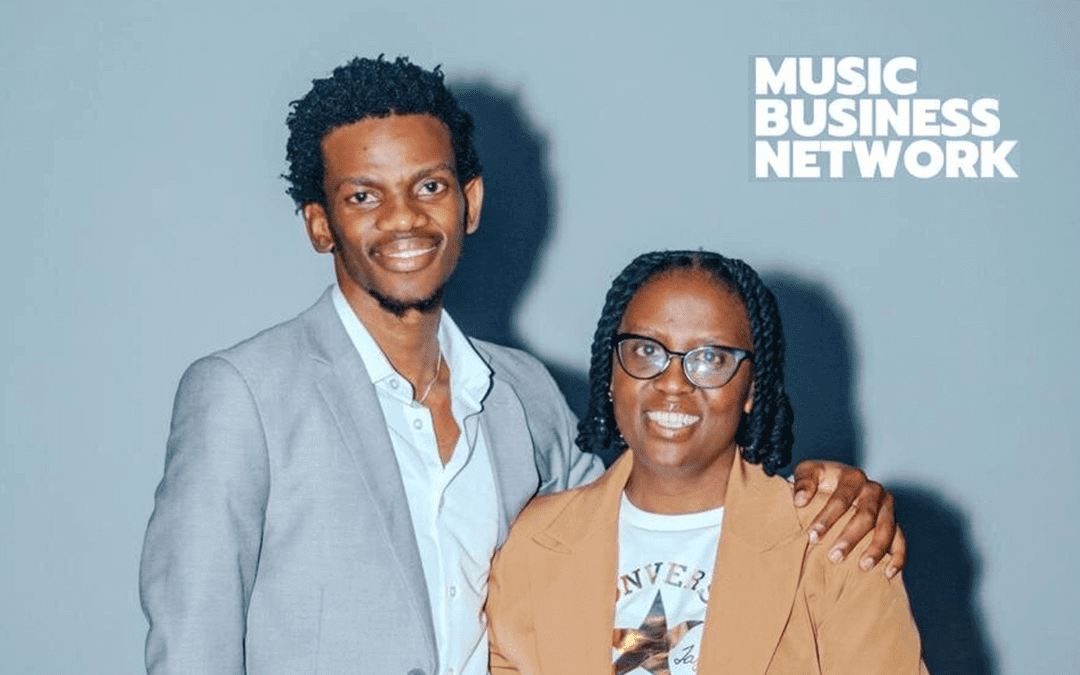 Two people stand side by side, smiling at the camera. The person on the left wears a light grey blazer and white shirt, with an arm around the other. The person on the right wears glasses and a tan blazer. “Music Business Network” text is in the top right corner.