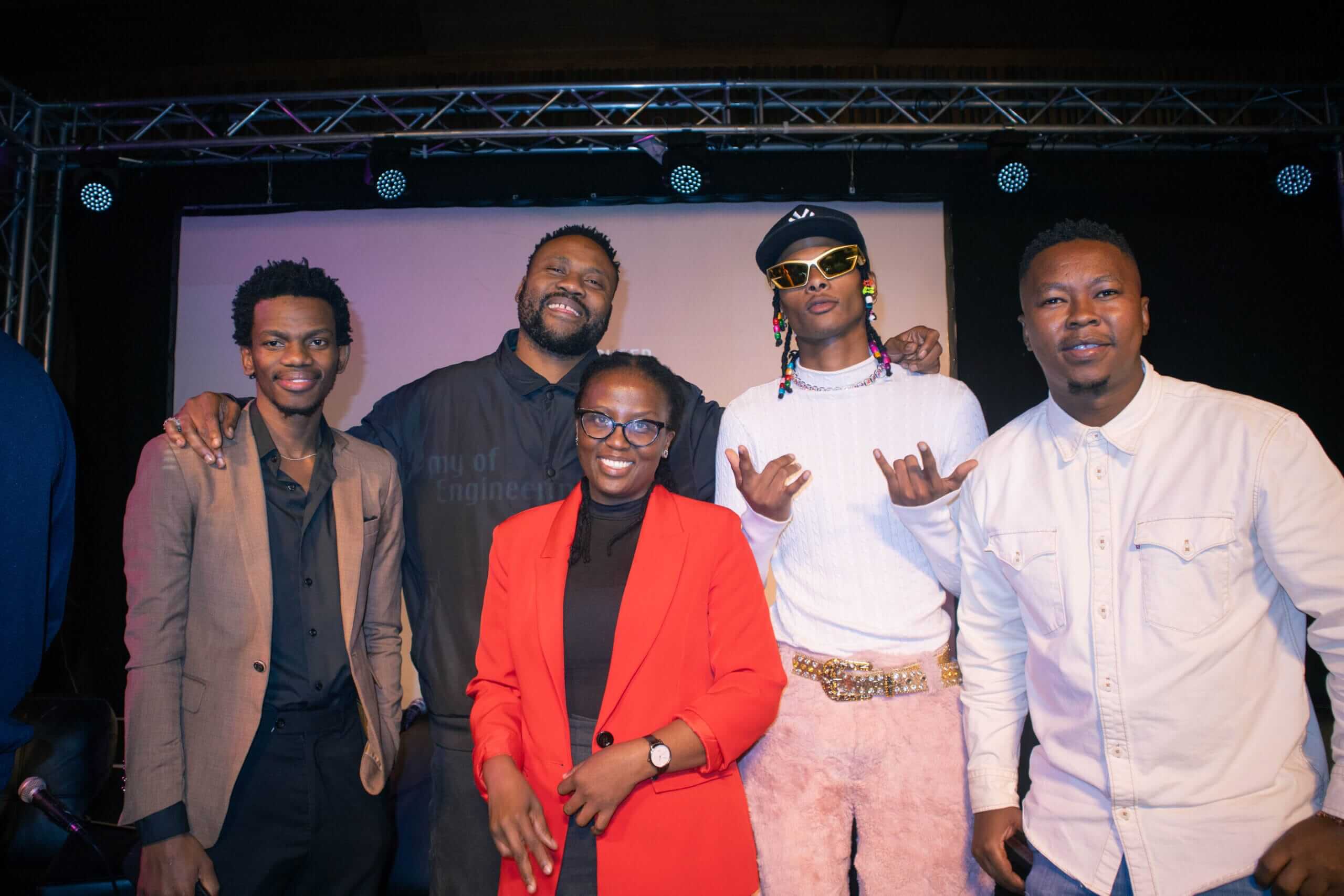 Five people pose together indoors in front of a stage. Three men stand on the left, one in a tan suit, one in a dark shirt, and one in a white jacket with sunglasses and colourful beads. A woman in a red blazer and glasses stands in front. They are all smiling or looking relaxed.