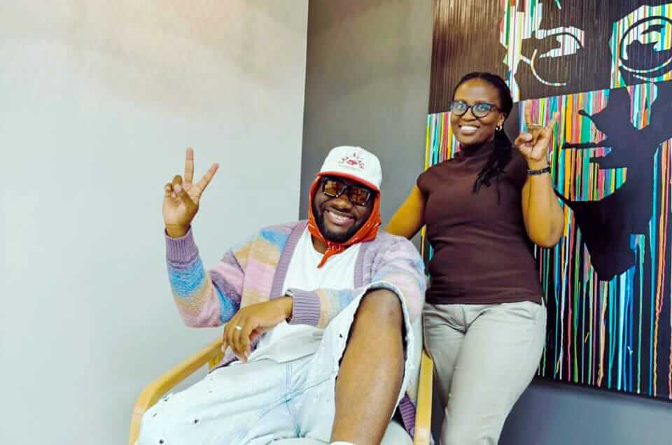 A smiling man in sunglasses and a colourful jumper sits on a chair making a peace sign, while a smiling woman stands beside him, also making a peace sign. Behind them is a vibrant, abstract artwork with faces and vertical coloured lines.