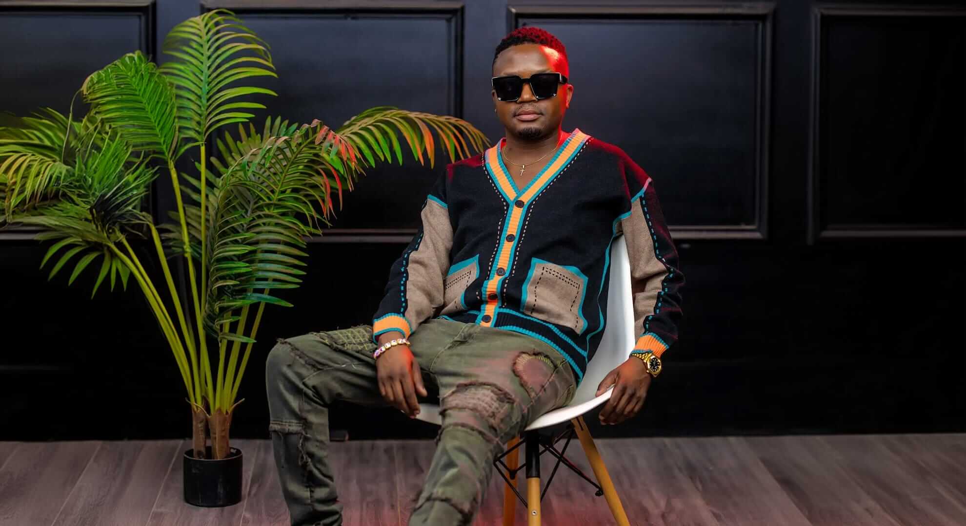 A man wearing sunglasses, a colourful cardigan, and ripped camouflage trousers sits confidently on a white chair. A large potted plant is beside him, and the background features black panelled walls and a wooden floor.