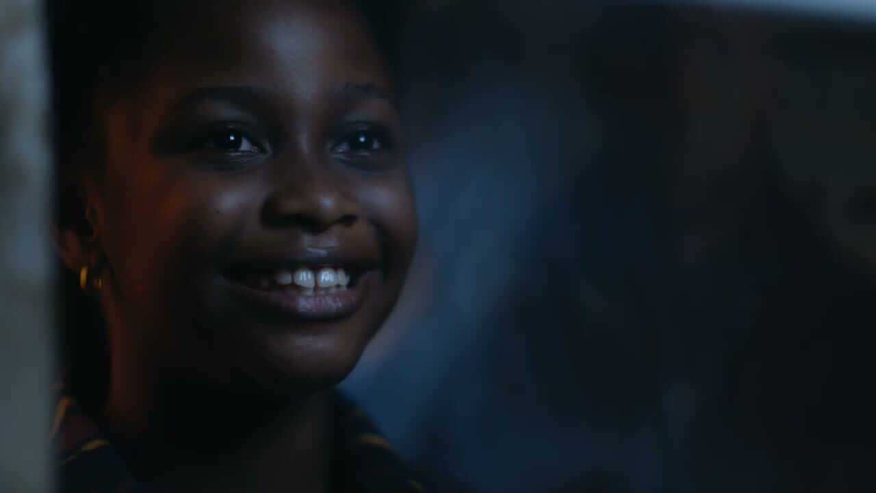 A young girl with dark skin smiles warmly in soft, dim lighting. Her face is partially illuminated, highlighting her bright teeth and sparkling eyes, whilst the background is blurred and dark, creating a gentle, intimate atmosphere.