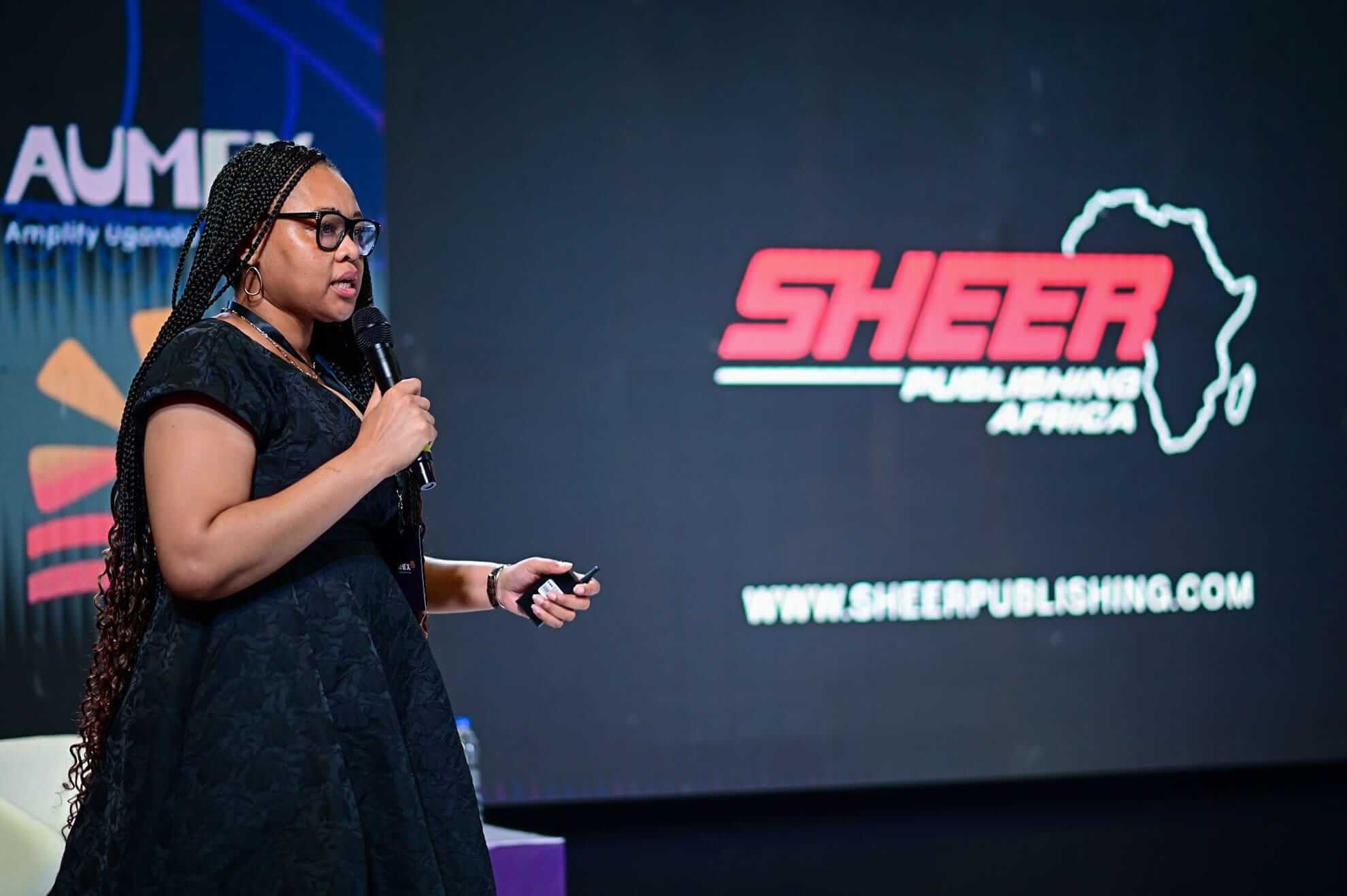 A woman with long plaits, wearing glasses and a black dress, speaks into a microphone on stage. Behind her is a large screen displaying the Sheer Publishing Africa logo and website, with a dark backdrop and colourful elements to the left.