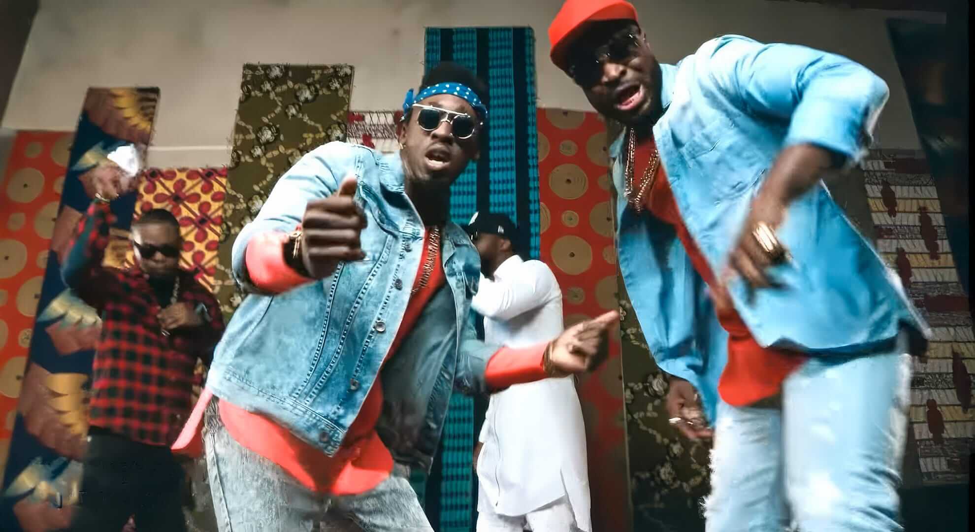 Four men dance energetically in a colourful room with bold patterned fabrics on the walls. Two men in front wear blue jackets, red shirts, sunglasses, and hats. The others in the background wear patterned and traditional outfits. The scene is lively and vibrant.