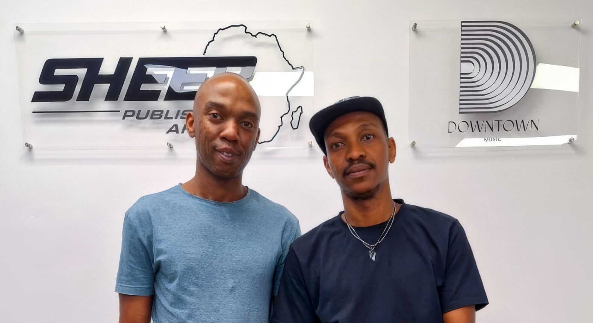 Two men stand side by side, smiling, in front of a white wall with two clear signs. The left sign has the SHEER Publishing Africa logo with an outline of Africa, and the right sign displays the Downtown Music logo with concentric circles.