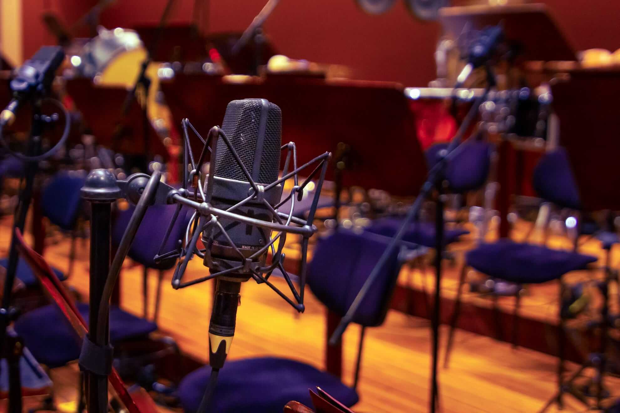 A close-up of a studio microphone with a pop filter in front of empty blue chairs and music stands in a warm-coloured room, suggesting a recording or rehearsal space for musicians, possibly an orchestra or ensemble.