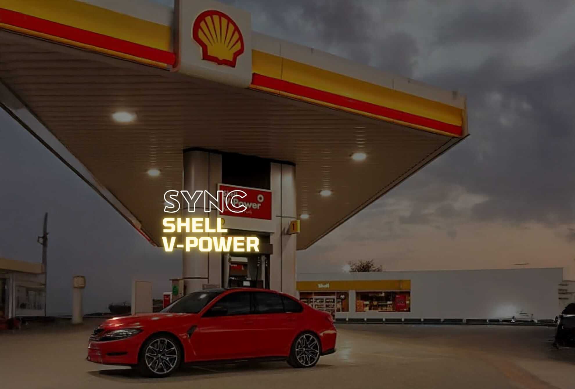 A red car is parked at a Shell petrol station at dusk. The station canopy and pumps are lit, with Shell’s yellow and red logo visible. A sign above the car reads 
