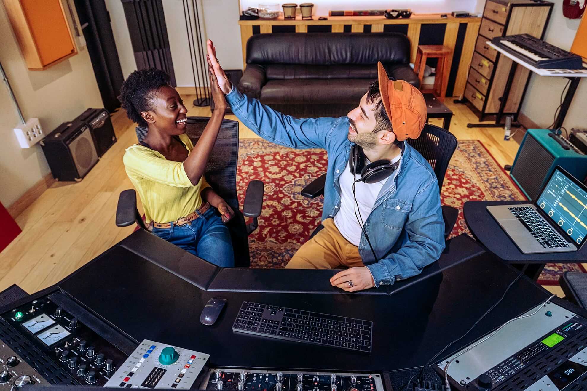 Two people sit in a music studio, smiling and giving each other a high five. One wears headphones and an orange cap, the other has short curly hair and a yellow shirt. They are surrounded by audio equipment, monitors, and a colourful rug.