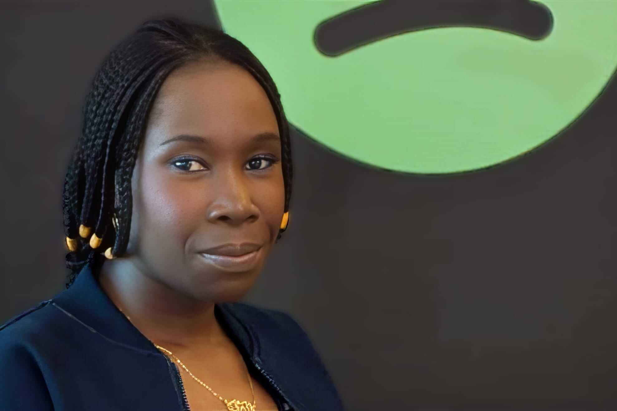 A woman with medium-dark skin and braided hair adorned with gold beads looks at the camera. She wears a navy jacket and a gold necklace. The background features a partial view of a large, green, abstract shape on a dark wall.