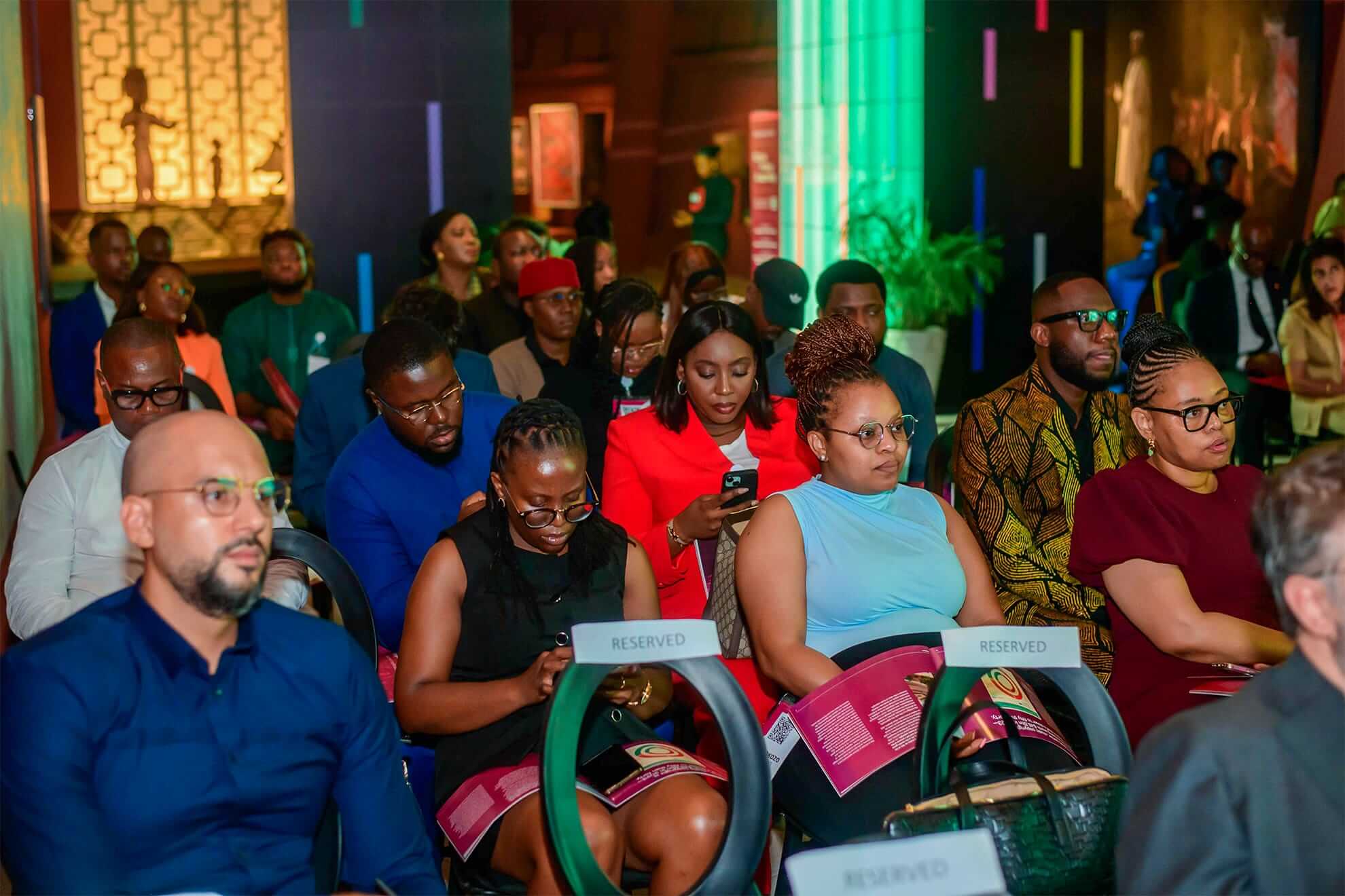 A diverse group of people sit closely together at an indoor event, some reading or using phones. The front row includes women in red and blue, with 