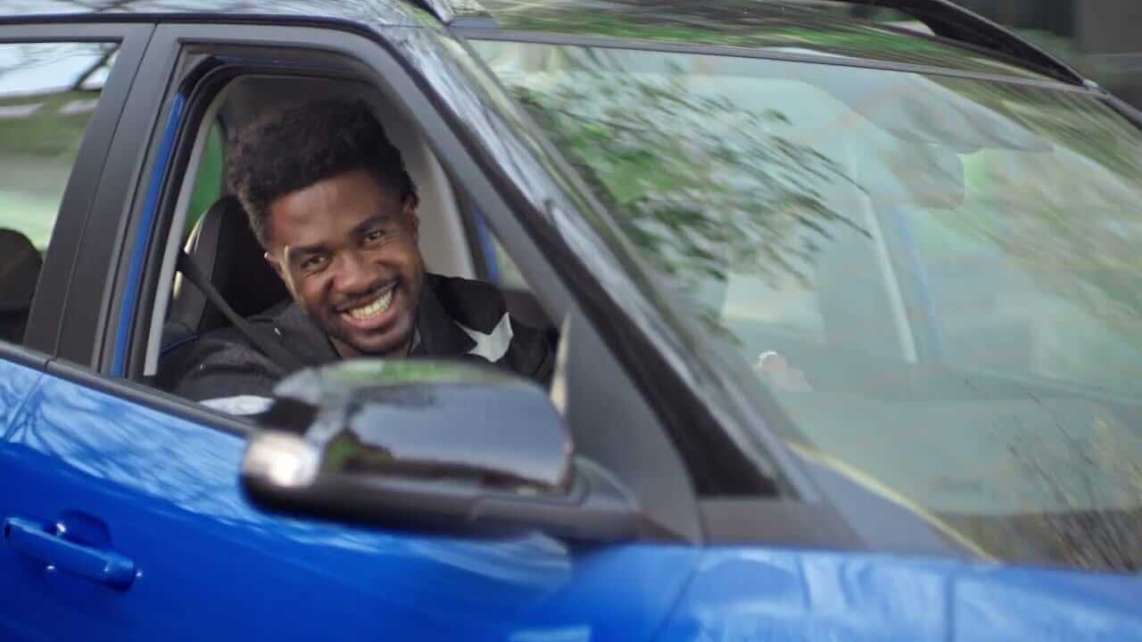 A young man with short curly hair smiles warmly while sitting in the driver’s seat of a blue car with the window down. Sunlight reflects on the car’s door and mirror, and trees are faintly visible outside in the background.