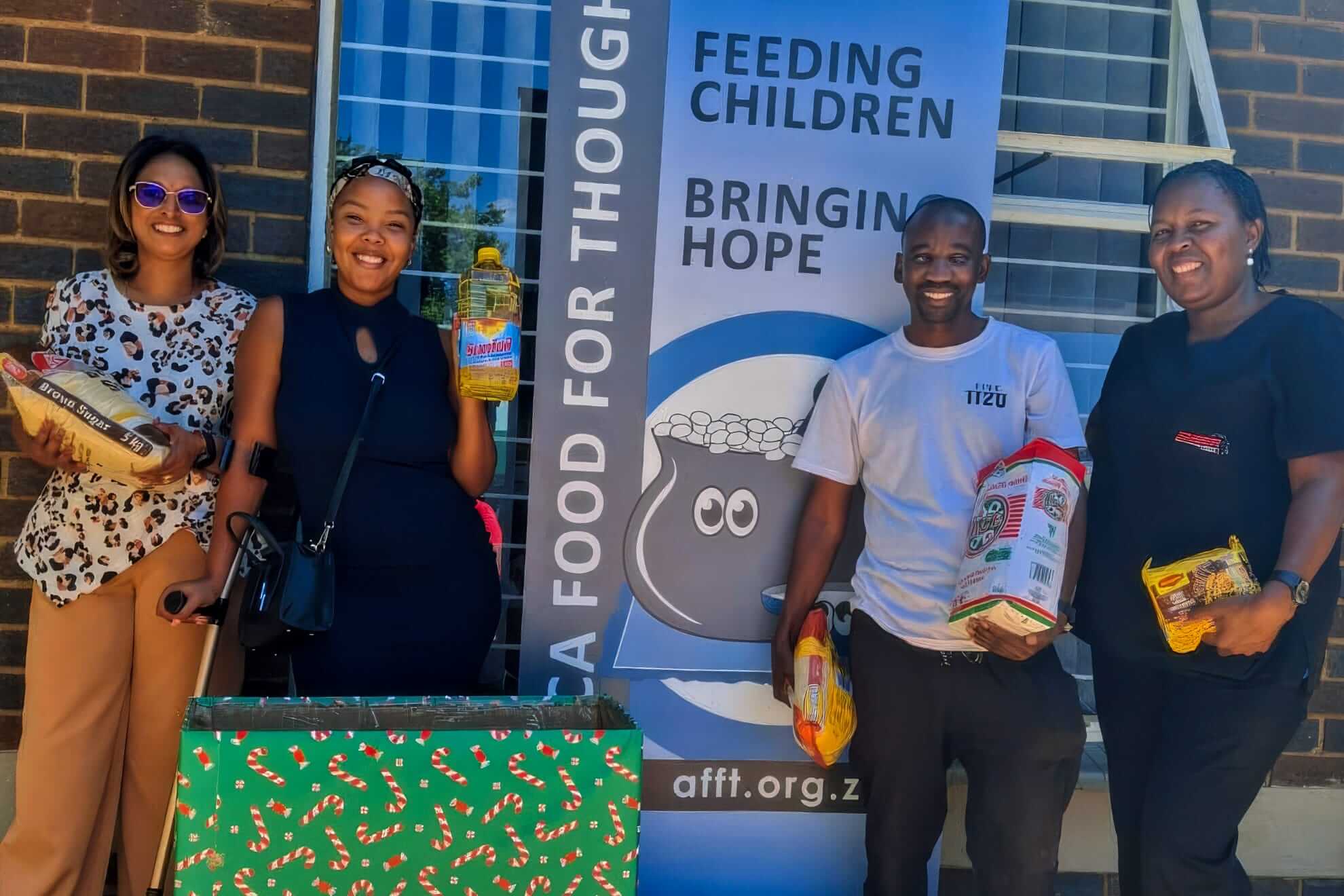 Four smiling adults stand outside by a brick wall, holding food items like oil and rice. Behind them is a blue banner reading, “Feeding Children, Bringing Hope” with a pot illustration and the website afft.org.za. A wrapped box sits in front.