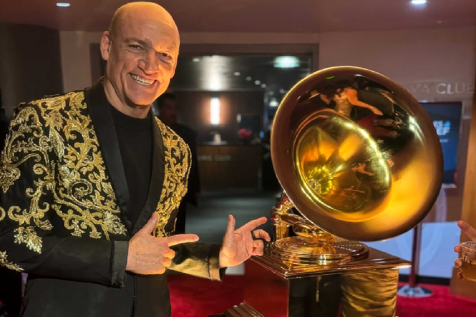 A bald man in a black suit with ornate gold embroidery smiles and points at a large golden Grammy award trophy. The background is softly lit, with red carpet flooring and people visible in reflections on the trophy’s surface.
