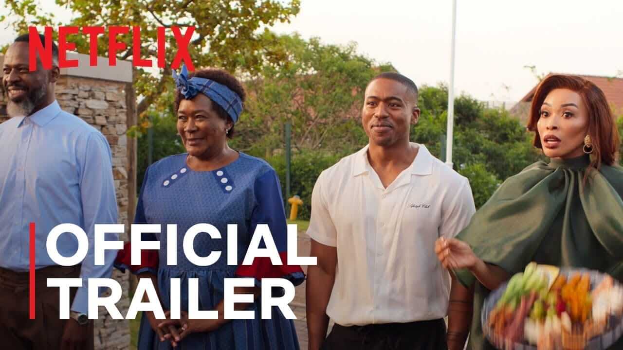 Four people stand outdoors, smiling. The man on the left wears a blue jacket, the woman next to him wears a blue dress and headband, the man in the middle wears a white shirt, and the woman on the right in green holds a colourful platter. 