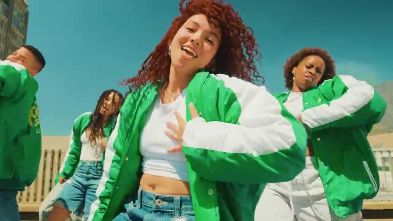 Four young people wearing green and white jackets dance energetically outdoors. The person at the front has curly red hair, a white crop top, and jeans. The background shows a clear blue sky and a building. Everyone looks joyful and expressive.