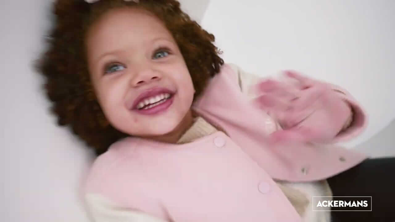 A young child with curly hair smiles joyfully while waving. The child wears a light pink coat over a cream sweater. The background is softly blurred in white tones. In the bottom right corner, the logo 