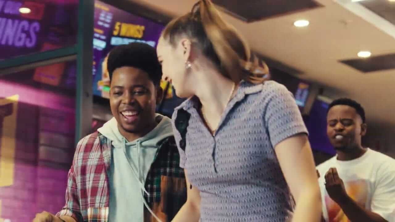 Three young adults enjoying themselves indoors, possibly at an arcade or entertainment venue. One person wears a plaid shirt and hoodie, smiling; another wears a purple knit shirt, laughing and dancing; and the third applauds in the background. Bright lights and screens are visible.