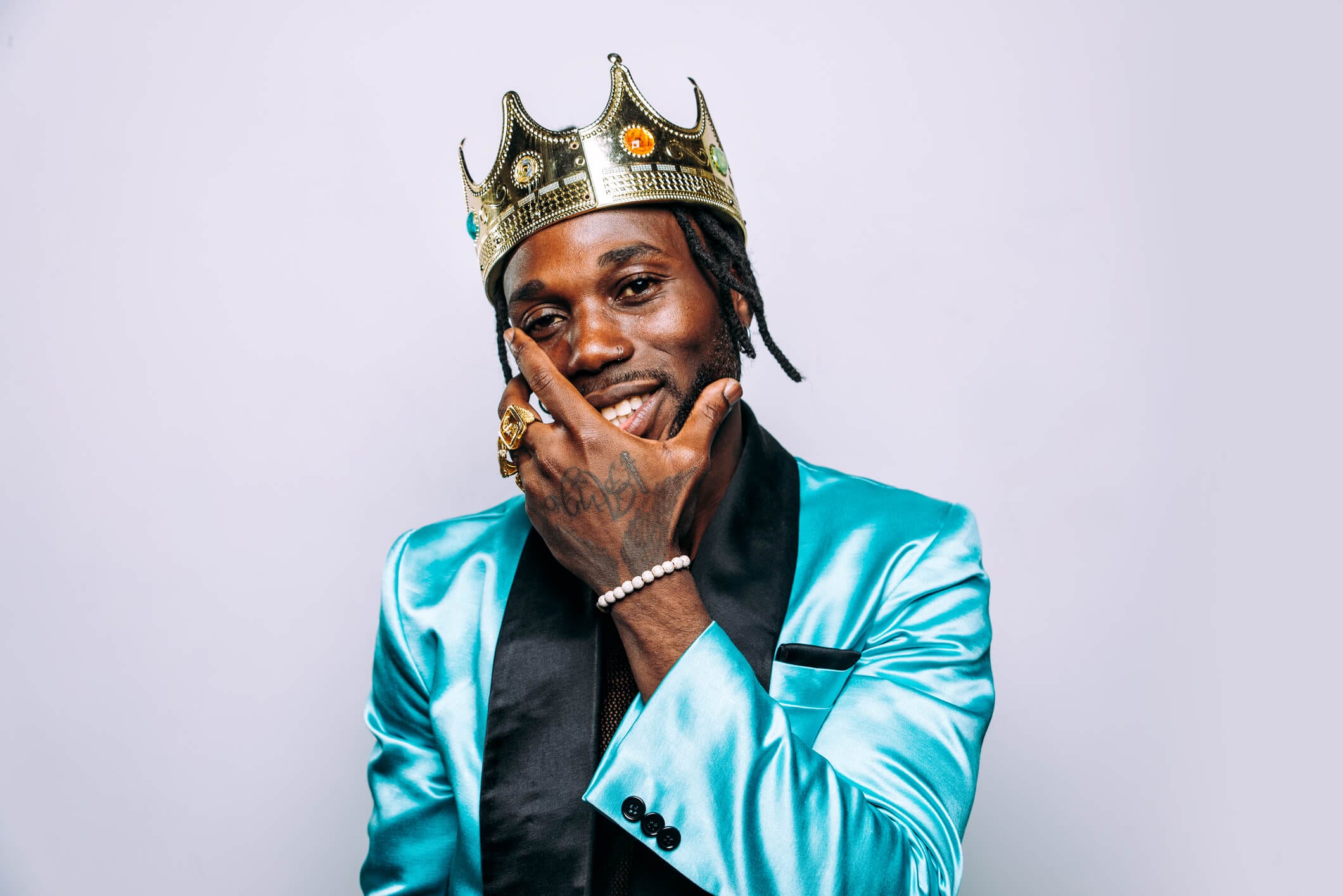A smiling man wearing a gold crown and a shiny turquoise suit jacket poses with one hand on his chin. He has rings, a beaded bracelet, and visible tattoos on his hand. The background is plain and light-coloured.