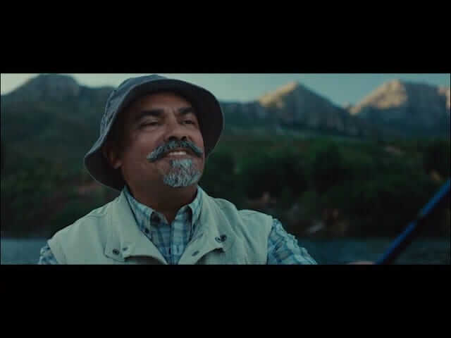 A middle-aged man with a gray goatee smiles while fishing outdoors. He wears a bucket hat, plaid shirt, and beige vest. Blurred green trees and rugged mountains are visible in the background under a blue sky.