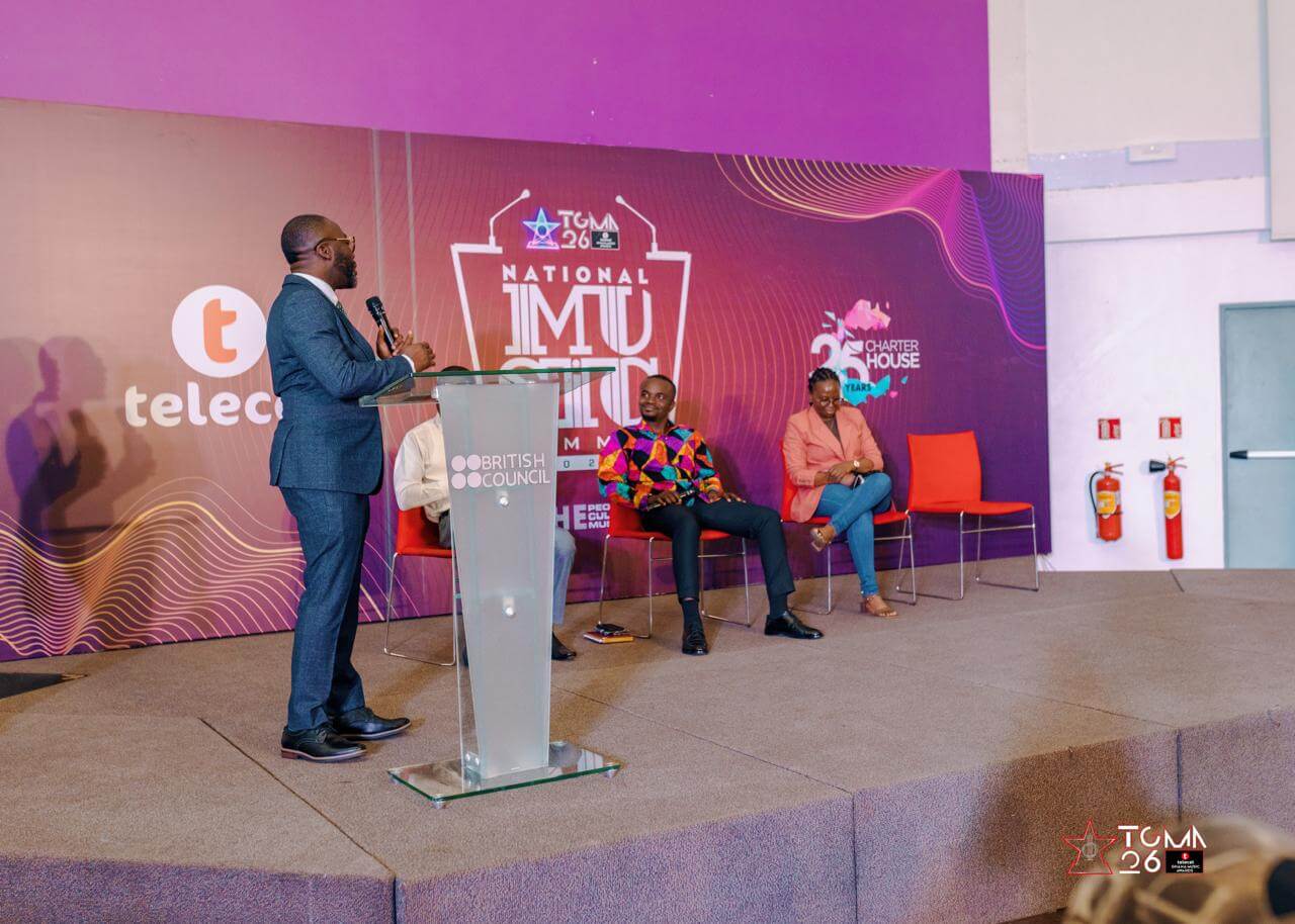 A man stands at a clear podium speaking to two seated men on a stage with a purple and pink backdrop labeled 