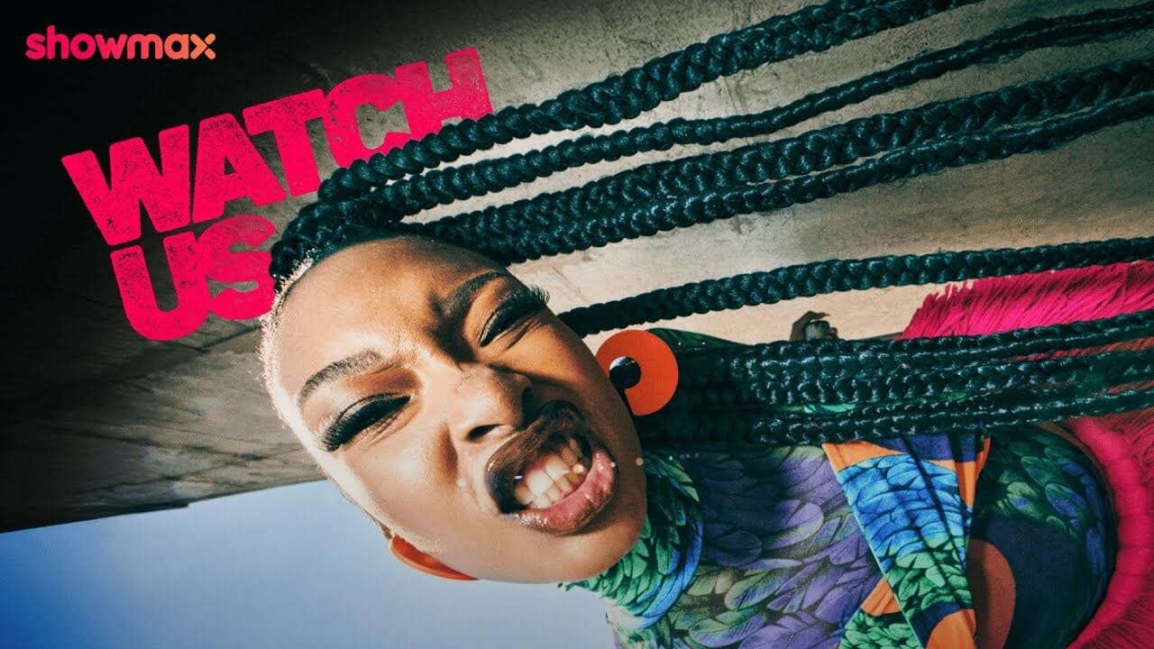 A woman with long braided hair and large orange earrings makes a fierce face at the camera. She wears a colorful, patterned top. Bold pink text reads 