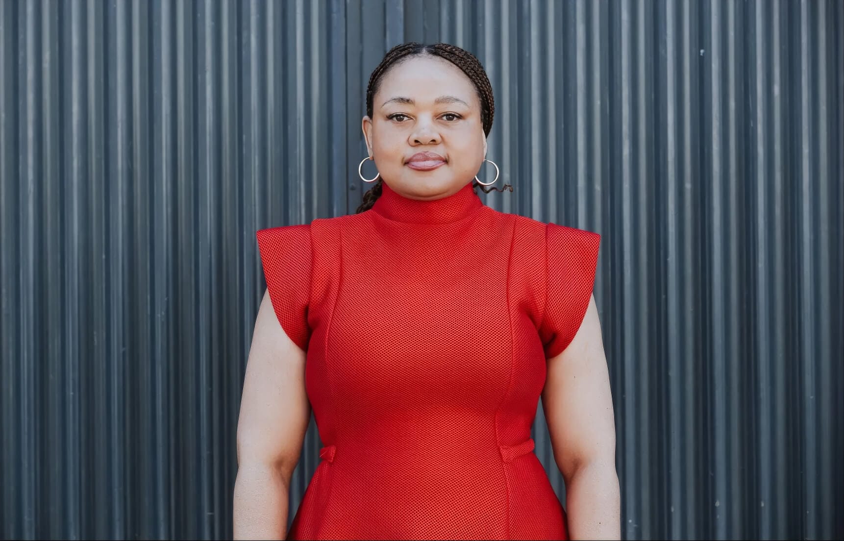 A woman with braided hair, wearing hoop earrings and a bright red, cap-sleeve, high-neck dress, stands facing the camera against a background of vertical black corrugated metal panels. She has a neutral expression.