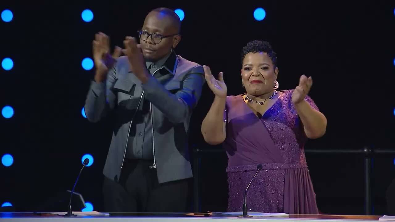 A man in glasses and a grey jacket and Nontokozo, in a purple, sparkly dress, stand side by side at a table, clapping. Behind them is a black background with rows of lit blue dots—both appear to be judges on Gospel Superstars.
