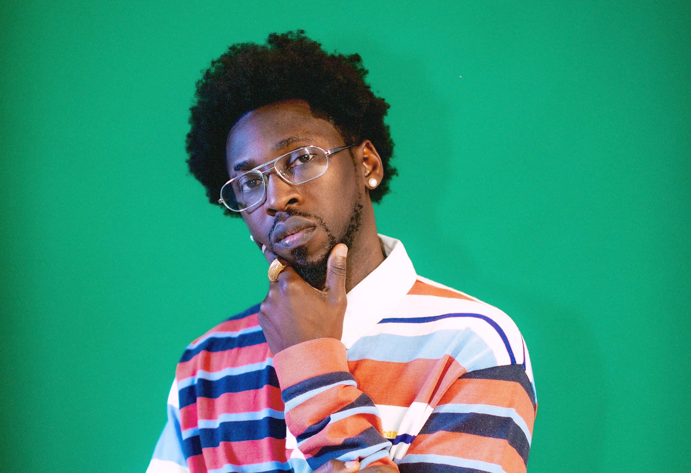 A man with short curly hair and glasses poses thoughtfully with his hand on his chin. He wears a colorful, striped long-sleeve shirt with red, blue, white, and tan stripes, and stands in front of a solid green background.