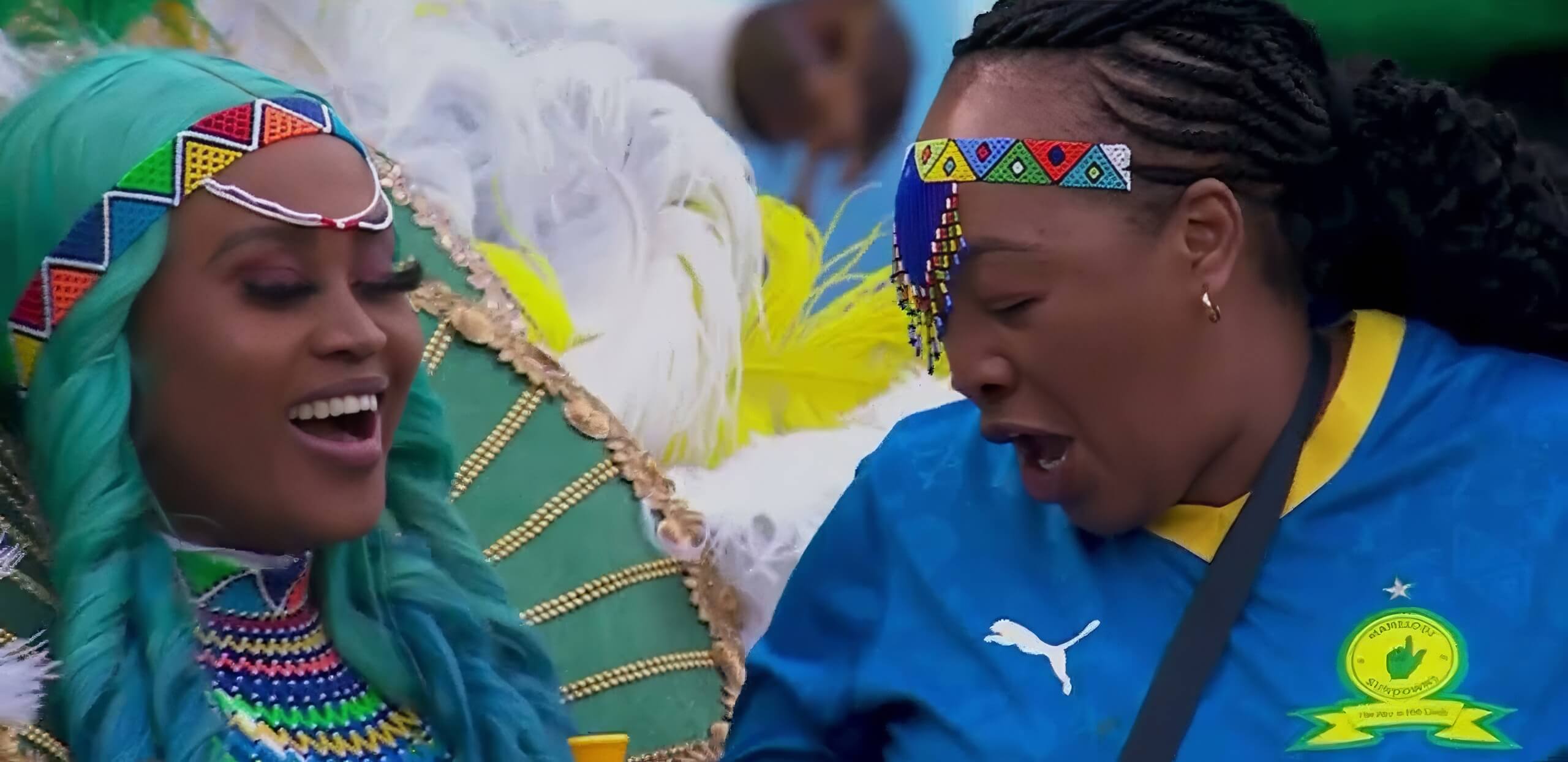 Two women joyfully celebrate. One wears a jewelled headpiece, teal wig, and vibrant feathered costume. The other wears a beaded headband and a blue Mamelodi Sundowns football shirt with a yellow collar and green trim. Both appear to be singing or cheering.
