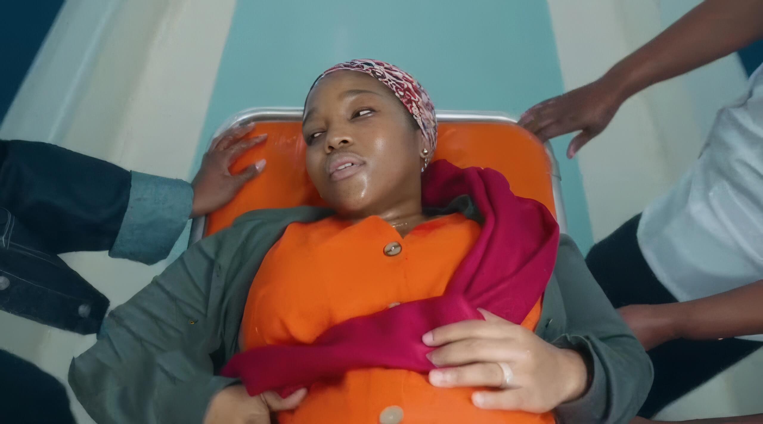 A woman in an orange dress and patterned headscarf lies on a trolley, looking distressed and sweaty. Two people’s hands gently support her as she clutches her stomach with one hand and a pink cloth rests on her torso. The scene suggests a medical emergency.