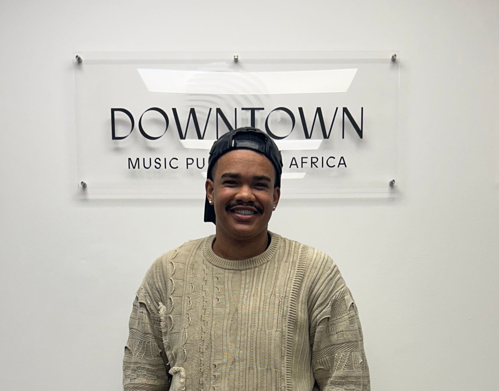 A person with a moustache and short hair, wearing a textured beige jumper and a backwards black cap, smiles whilst standing in front of a wall sign that reads 
