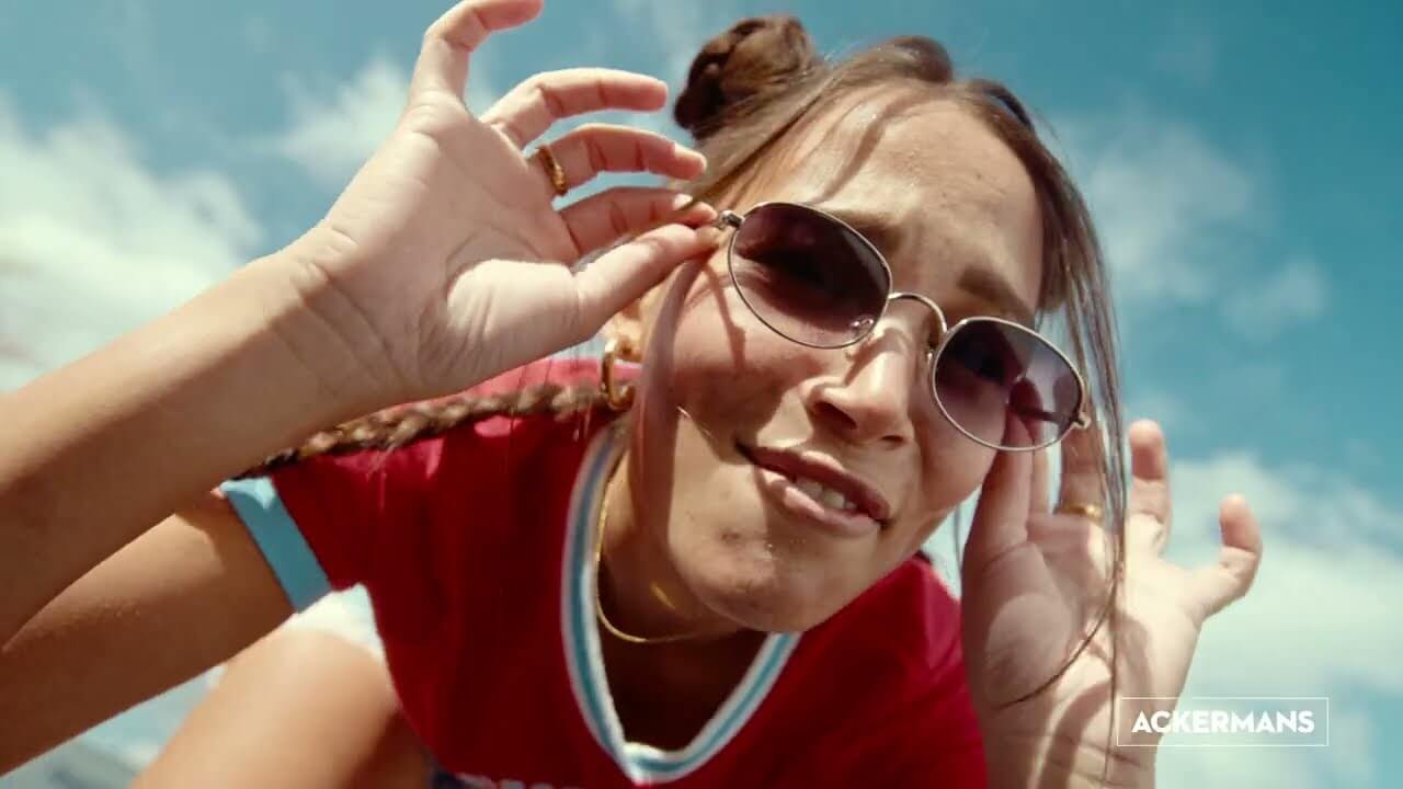 A young woman wearing round sunglasses, a red T-shirt, and gold rings leans towards the camera, adjusting her glasses with both hands. Her hair is styled in two buns, set against a blue sky. 