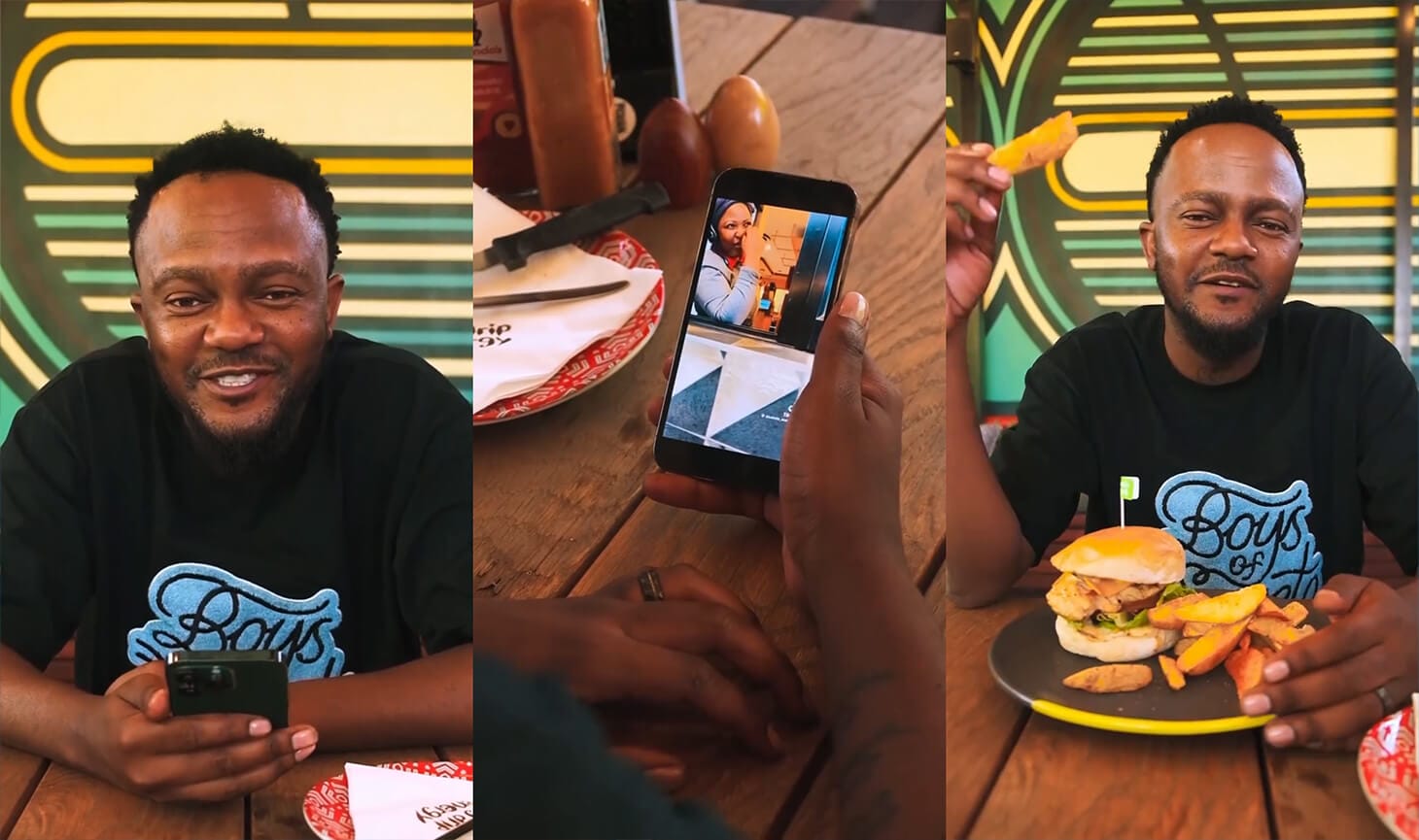 A man in a black “Boys” t-shirt sits at a wooden table in a colourful diner. He smiles, holds a mobile showing his photo, and eats chips next to a burger. Condiments and patterned decor are visible in the background.