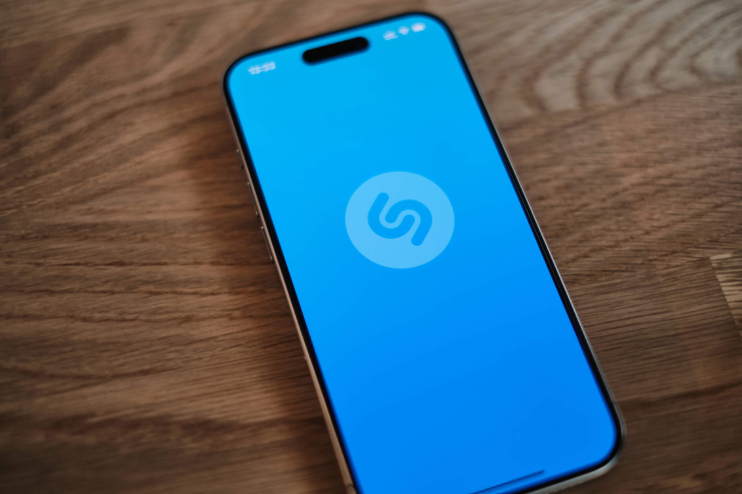 A smartphone rests on a wooden surface displaying the Shazam app’s bright blue splash screen with the white Shazam logo front and centre—perfect for discovering African music climbing the Global Top 200 charts.