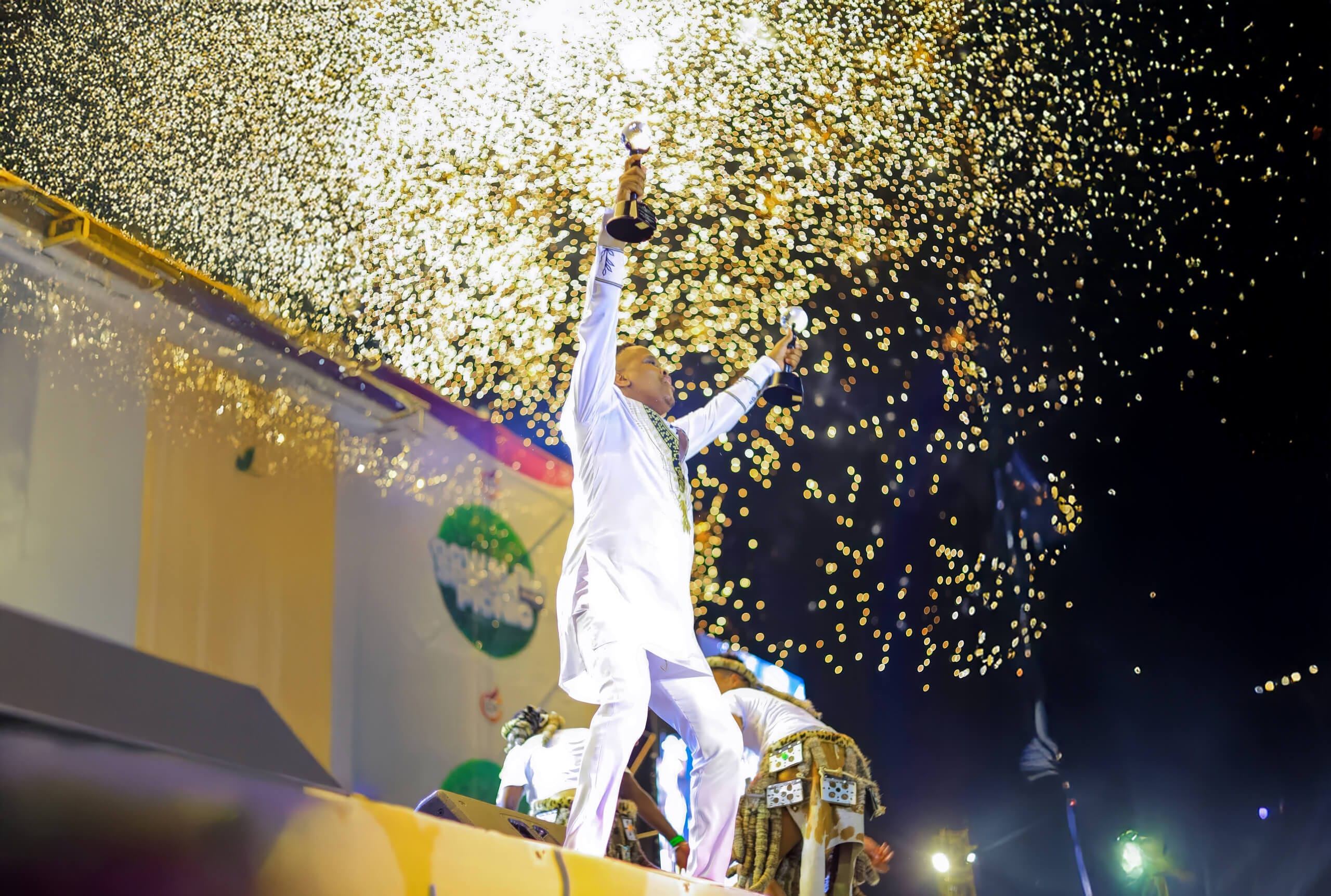 A person in white clothes stands on stage in Durban, holding a trophy aloft amid a burst of golden confetti and bright stage lights, celebrating Maskandi success at night with an enthusiastic crowd and other performers visible in the background.