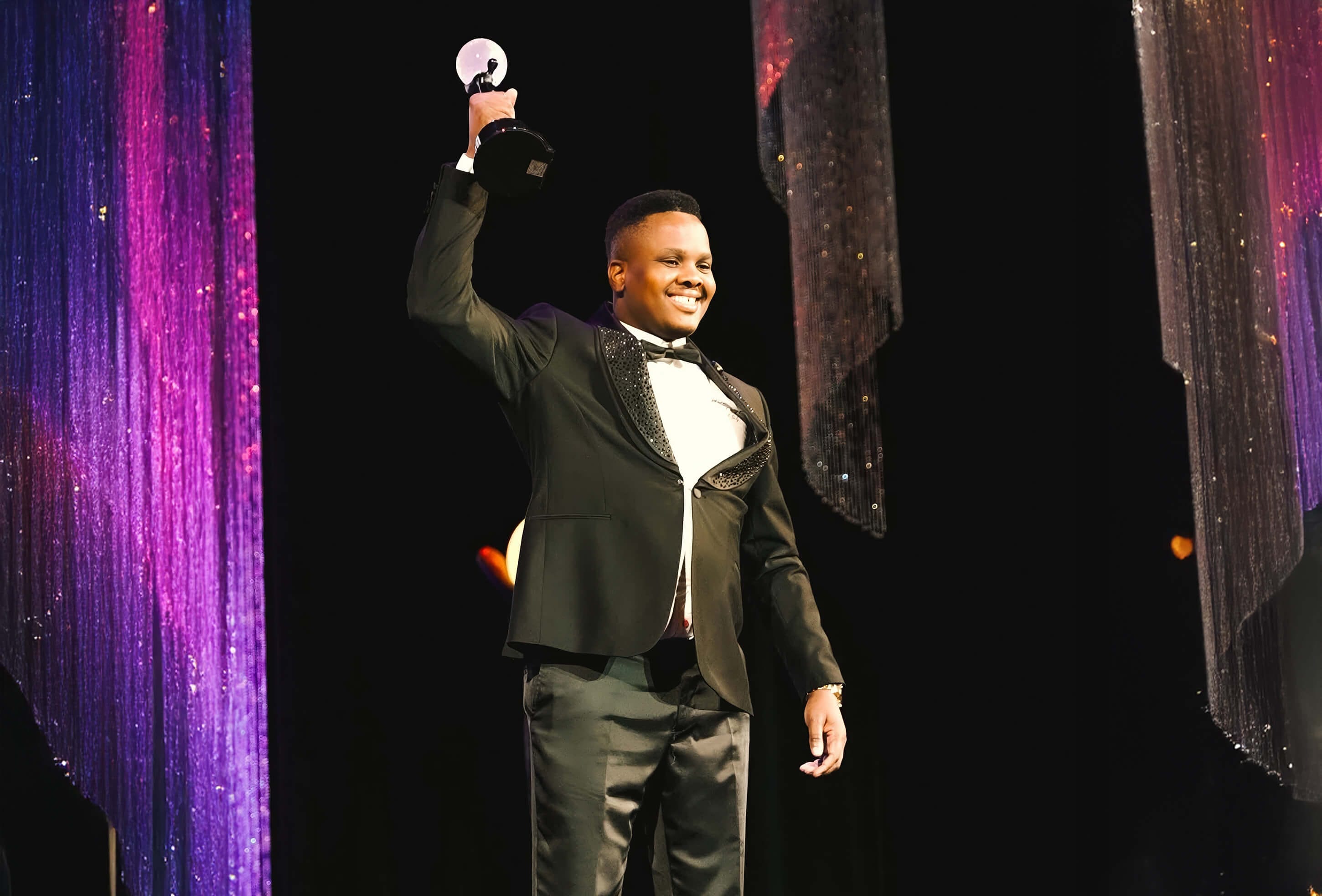 A man in a black tuxedo stands on stage, smiling and raising a trophy in one hand. Glittering purple, pink, and silver curtains frame the scene, creating a celebratory atmosphere. The stage lighting highlights his joyful expression and formal attire.