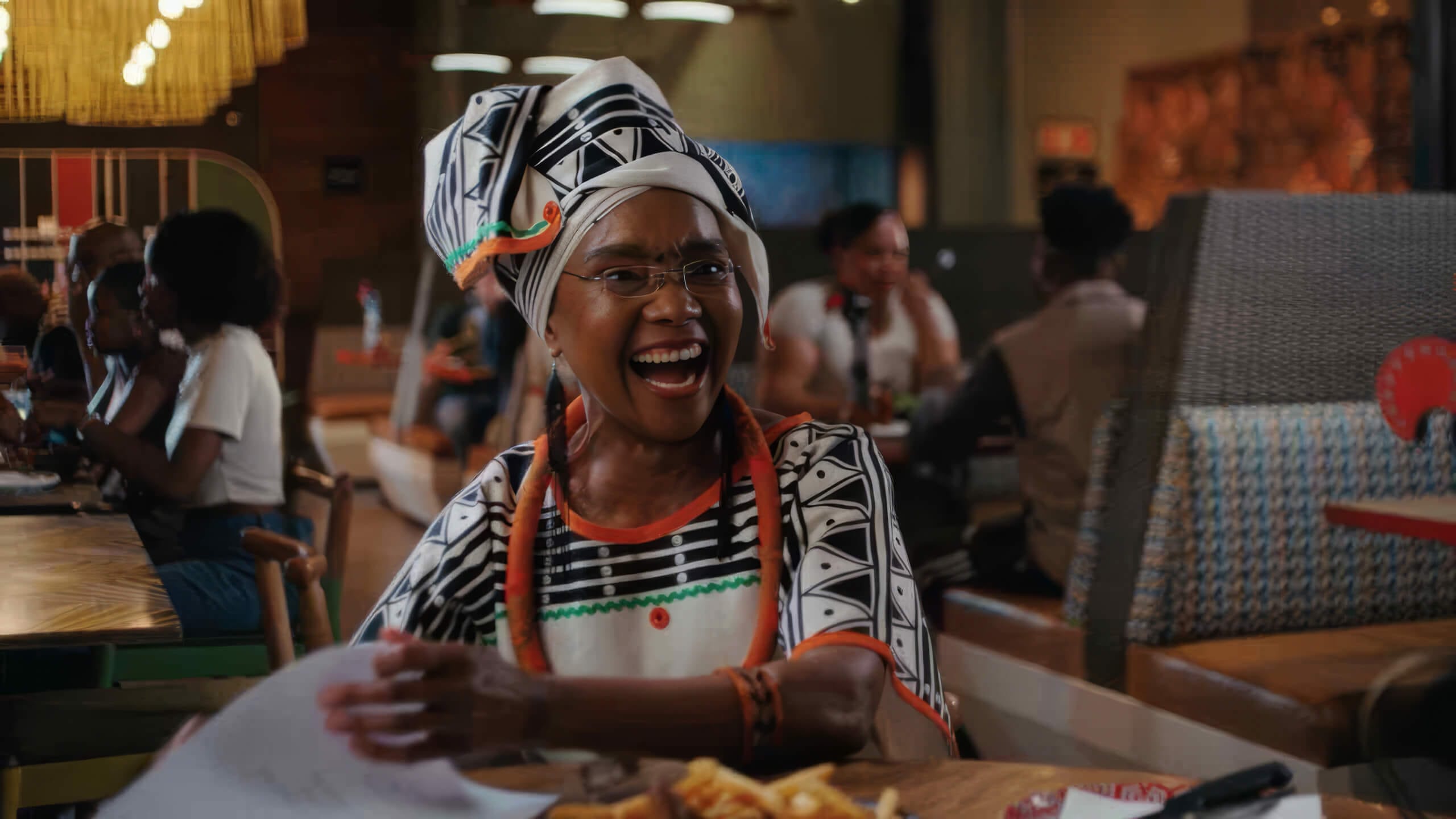 A smiling woman in traditional African attire and spectacles sits at a Nando’s South Africa table with food, holding papers and gesticulating animatedly. The colourful setting is lively, full of spirit, with joyful diners in the background.